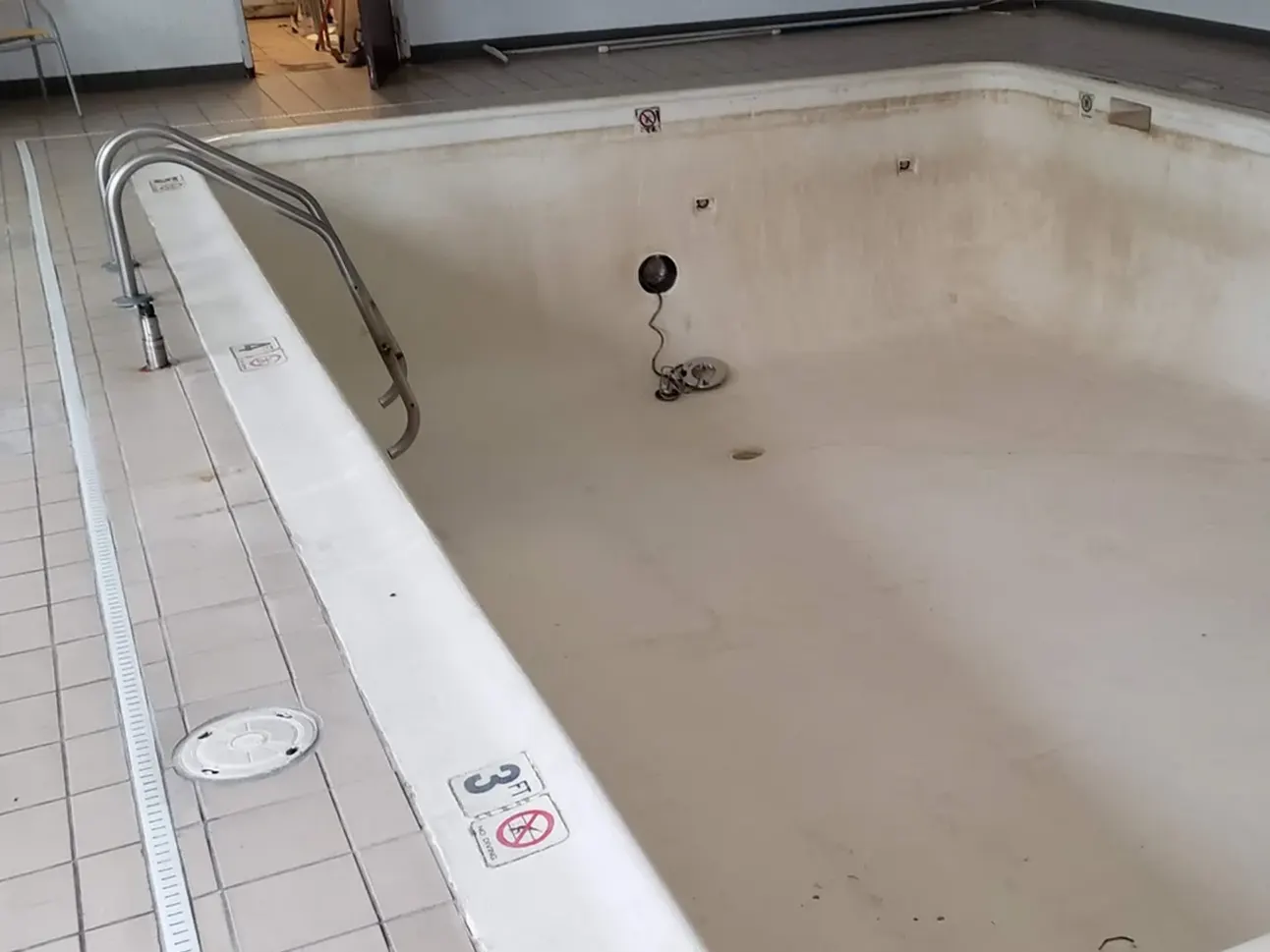 A hotel pool area before removal, showing a large, empty swimming pool with surrounding deck space and hotel buildings in the background. The pool is in need of demolition and repurposing.