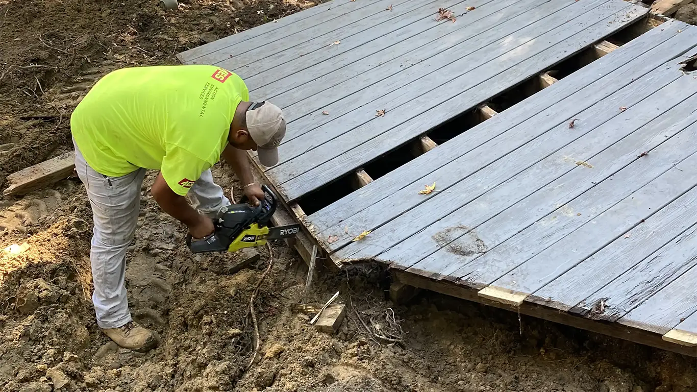 The image shows a worker in a bright yellow shirt using a Ryobi chainsaw to cut through a wooden deck as part of an above-ground pool removal. The deck is partially disassembled, revealing the underlying structure, with dirt and roots visible around the area. This highlights the process of above-ground pool removal services provided by Remove A Pool.