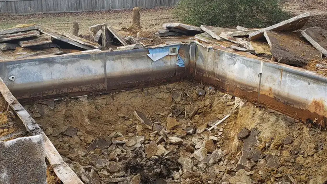 A partially demolished pool is filled with dirt and broken concrete as part of a pool removal in Allegany, MD. The rusted pool walls are still visible, with debris scattered around the site. The process appears to be in the midst of demolition, highlighting full pool abatement and removal services in the area, with landscaping services likely to follow.