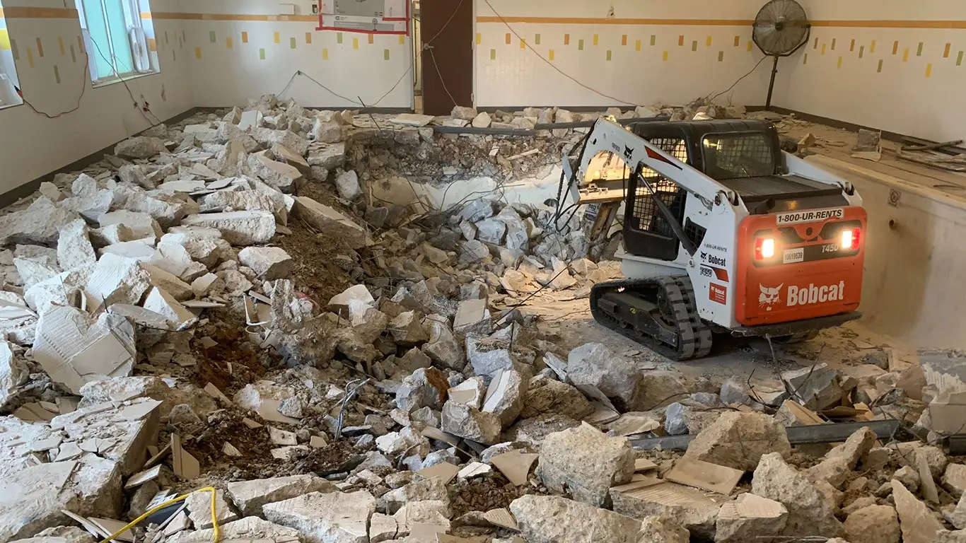 A Bobcat machine is actively demolishing an indoor pool, surrounded by piles of broken concrete and debris. The pool's walls have been broken apart, and the room's tiled walls and industrial fan can be seen in the background. This scene highlights the process of indoor pool removal by remove a pool across locations such as MD, VA, DE, PA, TX, and DC