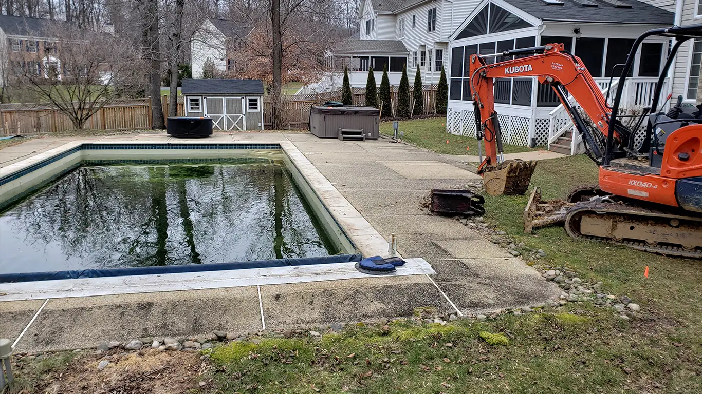 An inground gunite pool, filled with murky water, is set for removal as a Kubota excavator is positioned nearby, ready to begin the demolition. The pool is surrounded by a concrete patio, and a small shed and hot tub are visible in the background. This scene highlights the start of an inground pool removal process.