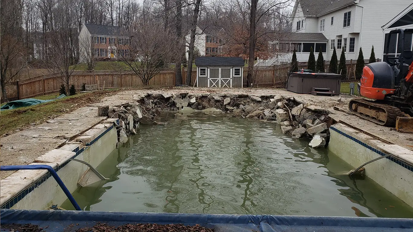 An inground pool partially demolished with broken concrete around the edges and water still filling the pool. An excavator sits on the right, with a backyard and shed in the background. Partial Pool Removal performed by Remove A Pool