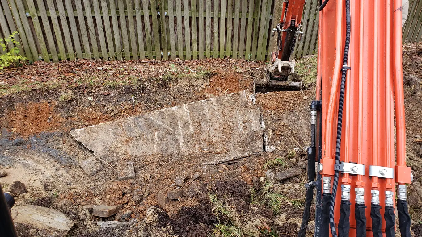 Heavy machinery is actively breaking apart a concrete slab as part of a pool removal in Florida. The excavator is lifting and cracking sections of the pool's foundation, with a wooden fence in the background, highlighting the demolition process for pool removal Florida.