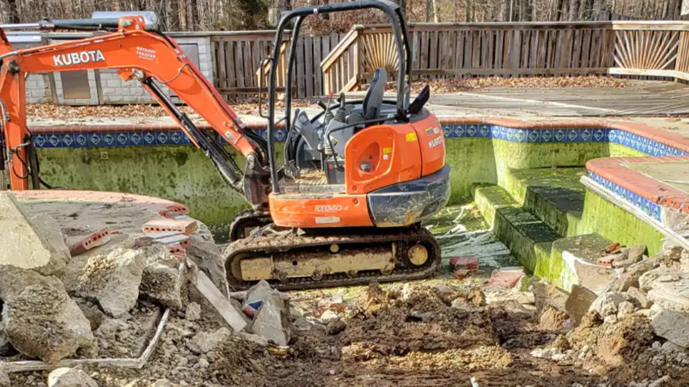 A Kubota excavator is demolishing an inground pool as part of a pool removal in Newark, DE. The pool, with stained green walls and broken concrete, is being dismantled, with debris scattered across the surrounding area. The adjacent patio and wooden fence are visible, highlighting the full pool abatement process, including pool removal and potential landscaping services.