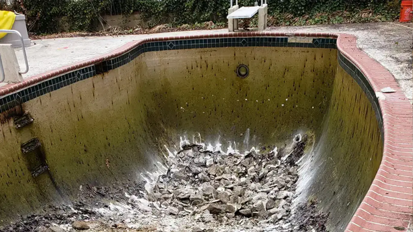 An empty, deteriorating pool is in the process of being demolished during a pool removal in Sussex, DE. The bottom of the pool is filled with broken concrete, and the sides are stained, showing years of wear. The red brick coping and remnants of a diving board stand above the pool, highlighting the beginning stages of the inground pool demolition.