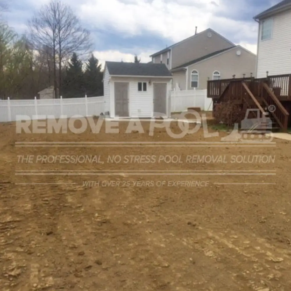 A backyard after a concrete pool removal by Remove A Pool. The ground has been leveled and cleared, with fresh soil visible where the pool used to be. A white shed and a wooden deck are visible in the background, surrounded by a white fence and trees.