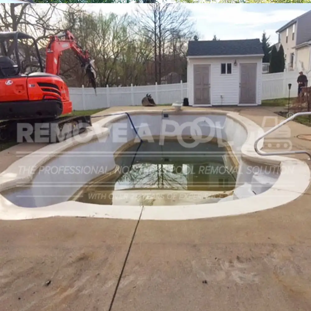 A backyard with a partially drained concrete pool before removal by Remove A Pool. An orange excavator is positioned near the pool, which is surrounded by a concrete deck. A white shed and a fenced yard are visible in the background.