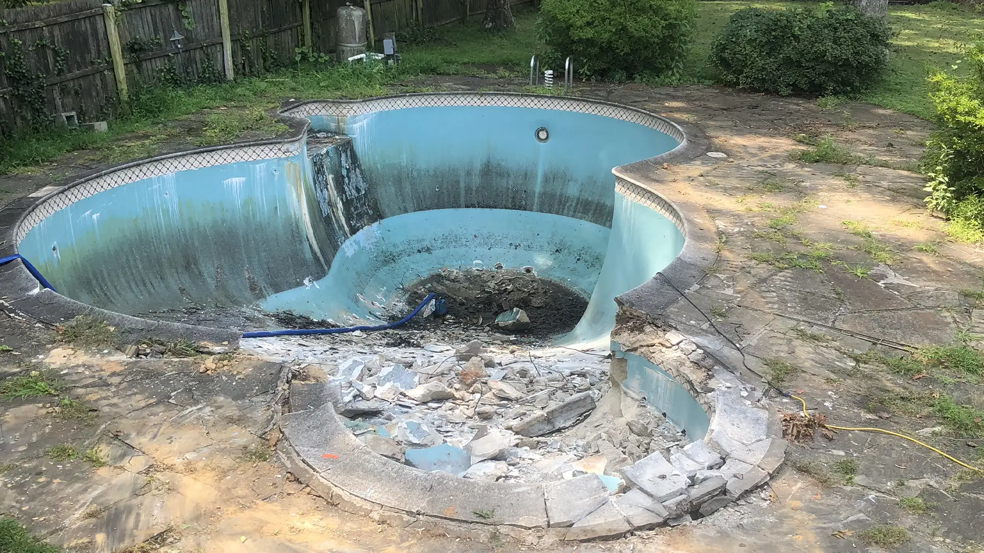 An in-ground swimming pool undergoing demolition as part of a full pool abatement service by Remove A Pool. The pool’s interior is partially demolished, with broken concrete and debris scattered at one end. The pool's blue surface is stained, and the surrounding concrete deck shows signs of wear. A wooden fence and overgrown vegetation frame the yard in the background.