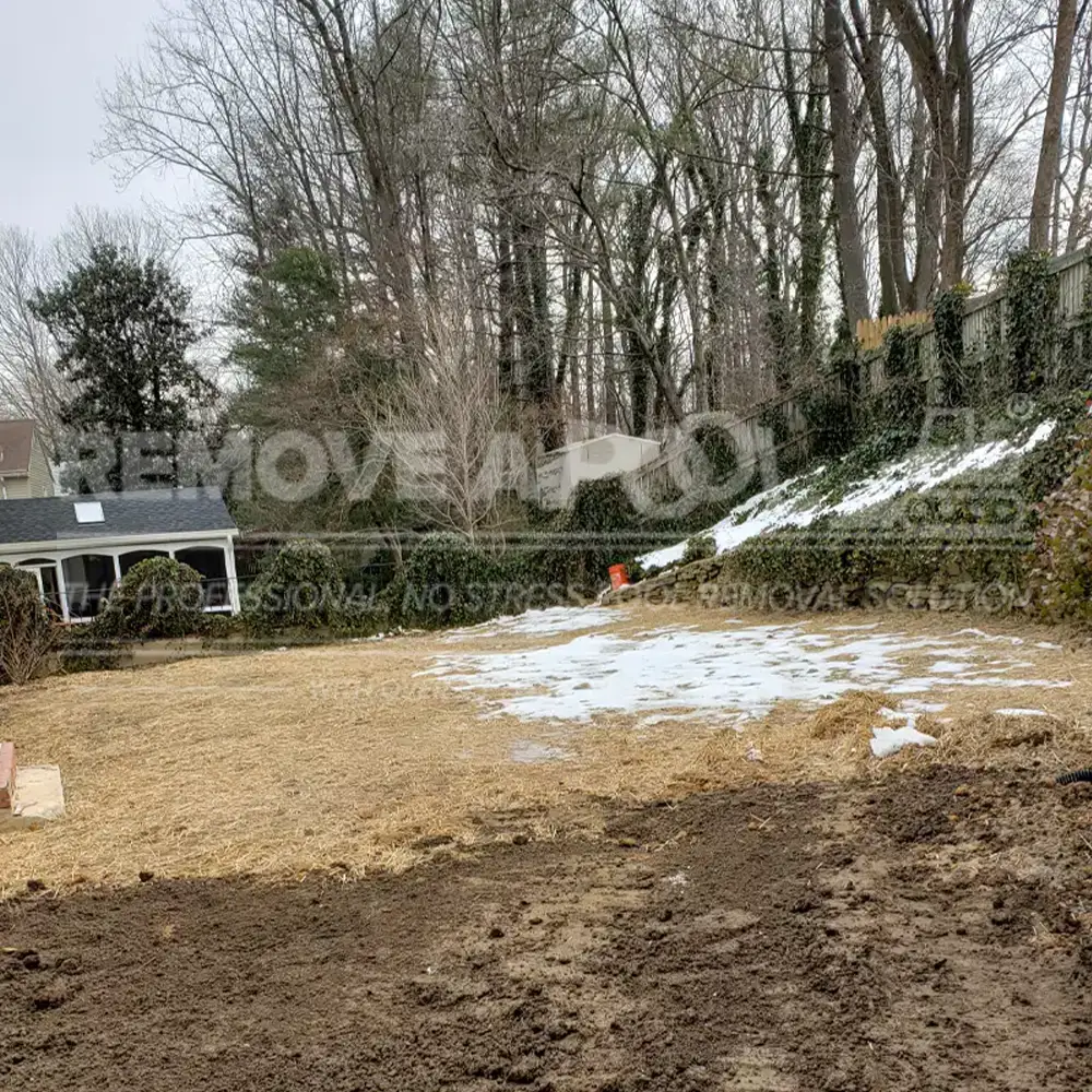 A yard after a pool removal by Remove A Pool, with the ground covered in straw and some remaining snow. The area is enclosed by ivy-covered fences and trees, with a small shed and a house in the background.