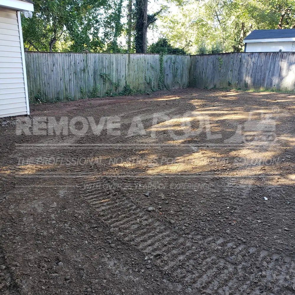 A backyard after the removal of a gunite pool by Remove A Pool. The ground has been leveled and cleared, with visible tracks from heavy machinery. A wooden fence encloses the yard, and a house is partially visible on the left side.