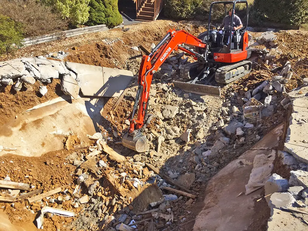 A mini excavator demolishing a concrete in-ground swimming pool, with the surrounding area filled with rubble and broken concrete pieces. The operator is seated in the machine as it clears out debris from the site.