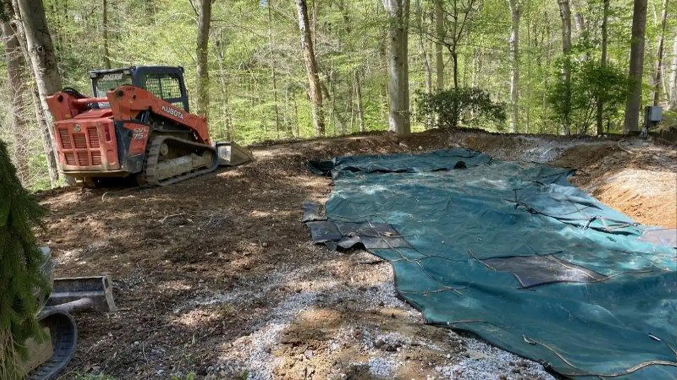 The image shows a backyard pool removal in progress in Delaware. A small red Kubota construction vehicle is parked near a forested area, while a large green tarp covers the ground where a pool once was. The tarp protects the excavation site, with trees and loose soil surrounding the area, highlighting the pool removal Delaware project.