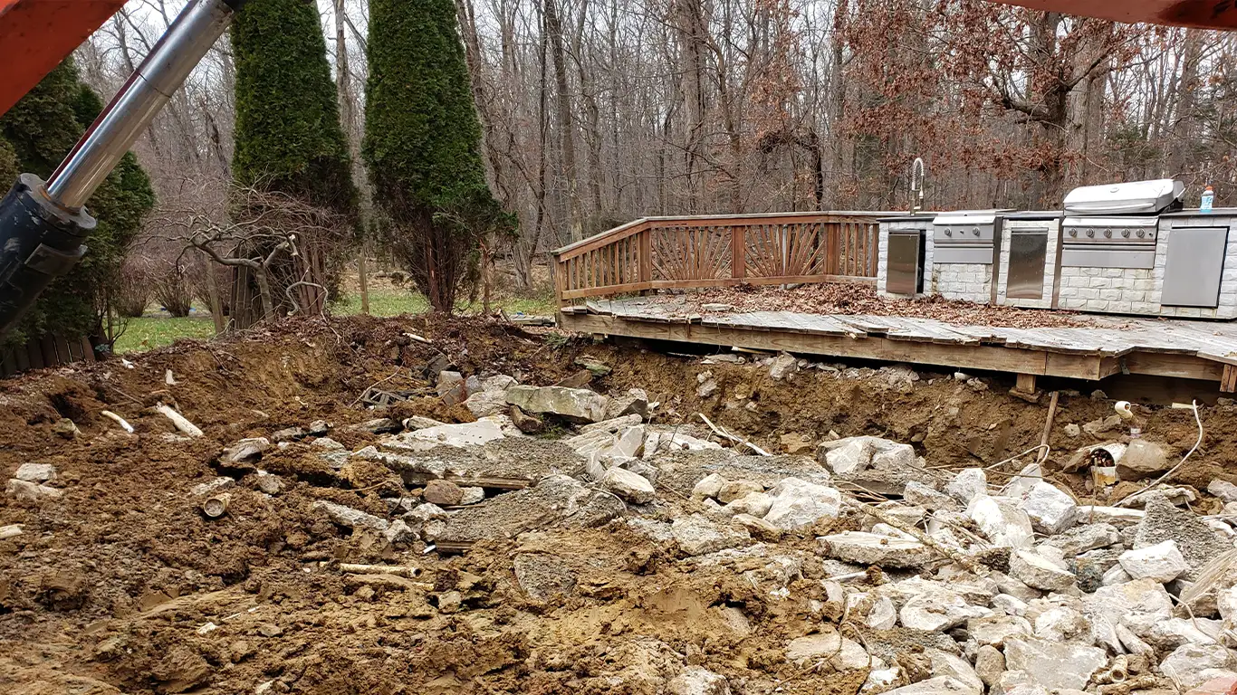 The image shows a backyard pool removal in progress in Dover, DE. The pool area is filled with broken concrete and soil, while an outdoor kitchen and patio deck are visible in the background. Heavy machinery is working on the site, with large piles of debris scattered throughout the excavation zone. This highlights the detailed process of pool removal Dover DE.
