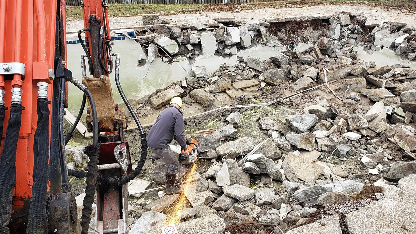 The image shows a worker using a saw to cut through large concrete pieces during a pool removal in Kent County, DE. An excavator is visible in the background, surrounded by piles of broken concrete as the pool is being demolished. The scene captures the detailed process of pool removal Kent County DE, with heavy machinery and manual labor involved.