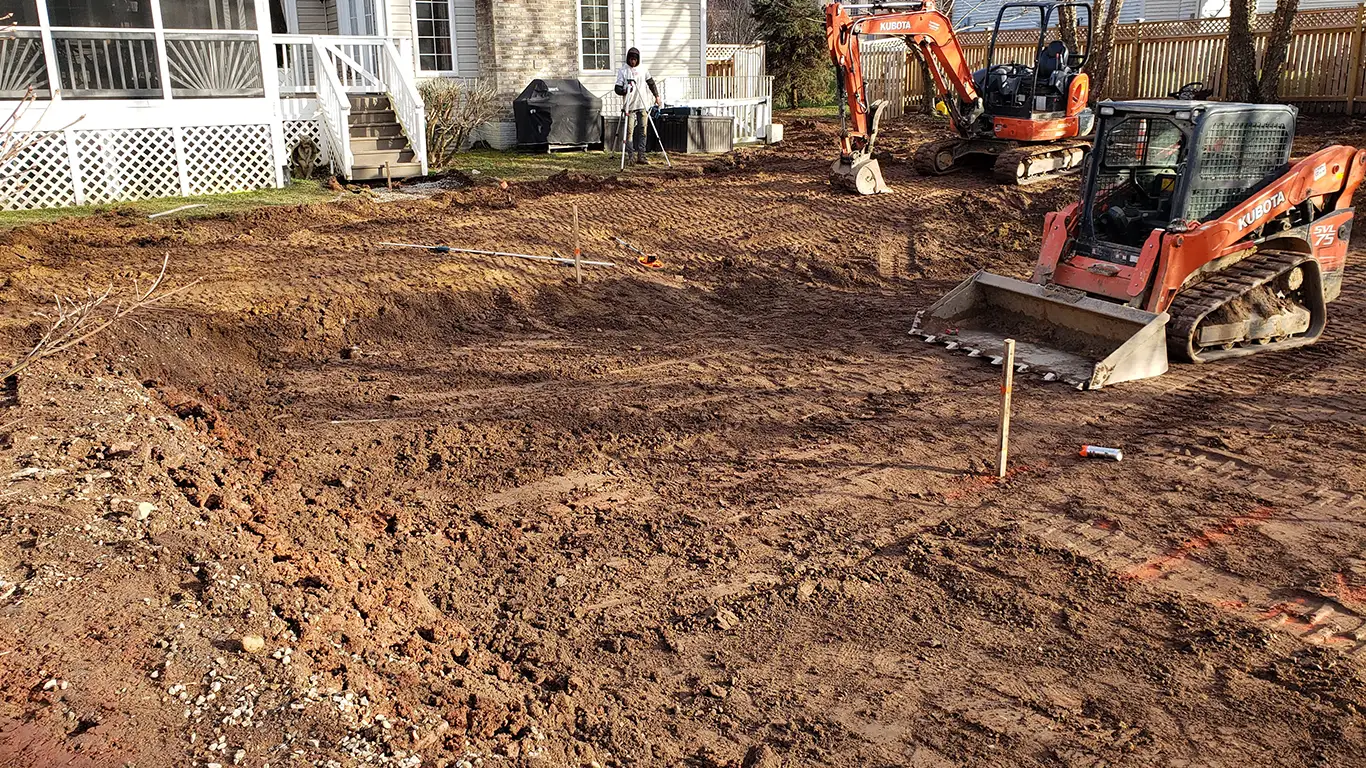 The image shows a completed pool removal in Lewes, DE, with two Kubota excavators parked on the leveled ground where the pool used to be. The area has been backfilled and smoothed over, while workers stand nearby, taking final measurements. The project appears to be nearing completion, emphasizing the pool removal Lewes DE process.