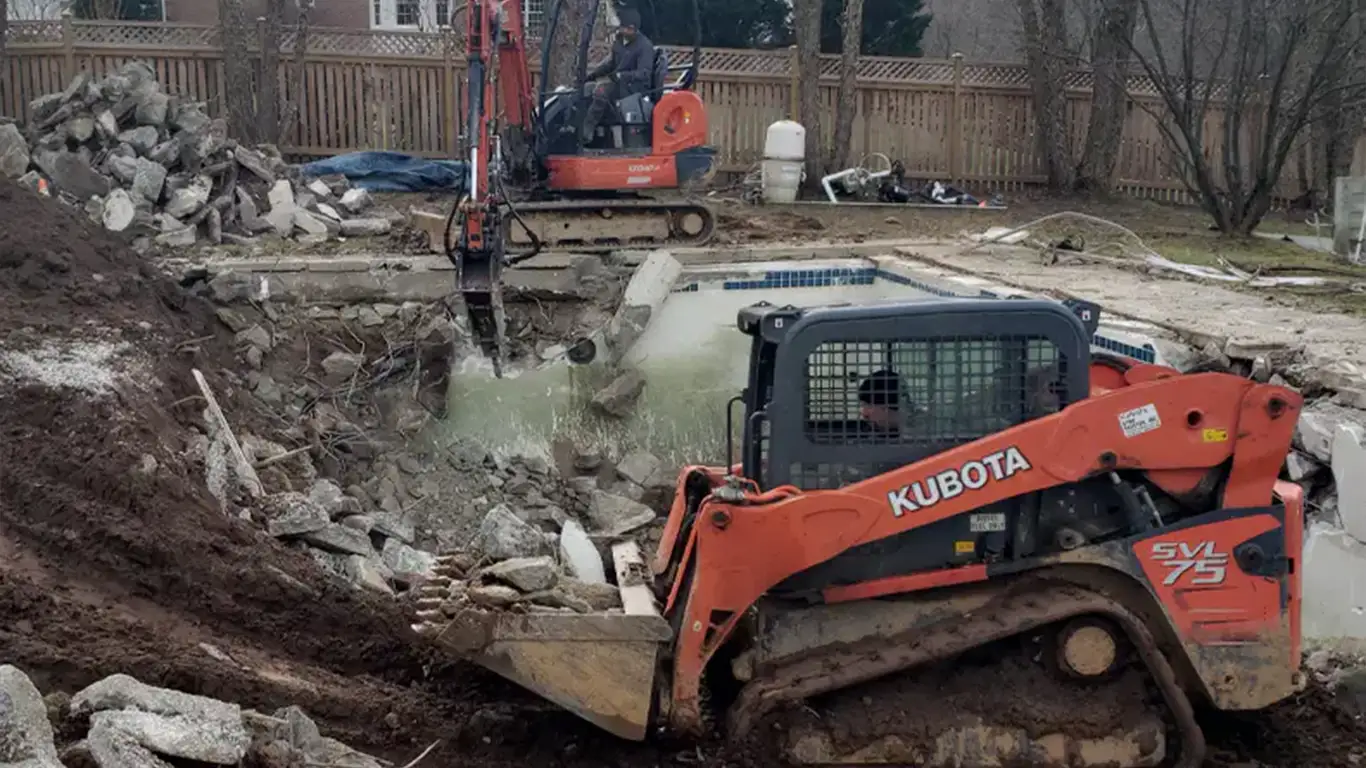 The image shows a pool removal Wilmington, DE. A Kubota SVL75 skid steer is working alongside another excavator to break up and remove a concrete in-ground pool. Debris is scattered around the pool area, and large piles of broken concrete are visible, highlighting the extensive demolition required for the pool removal process. The backyard is bordered by a wooden fence, with some equipment and materials seen in the background.