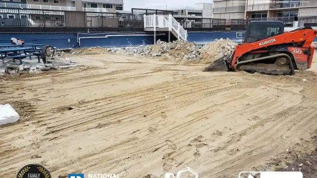 An excavator leveling the sand and soil at the former pool site, leaving smooth tracks across the surface.